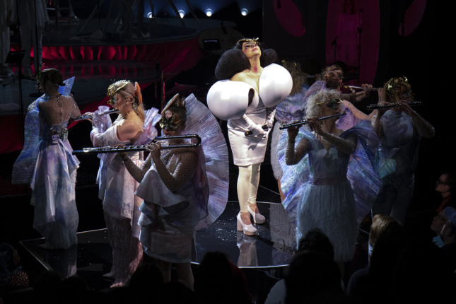 Björk stands center in a white dress with oversized shoulders while flautists, in white dresses with wing-like backs, play around her. They all wear masks.