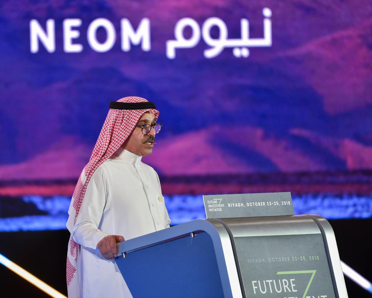 一名男子在讲台上讲话，他身后有紫色的标志，上面用英语和阿拉伯语写着NEOM