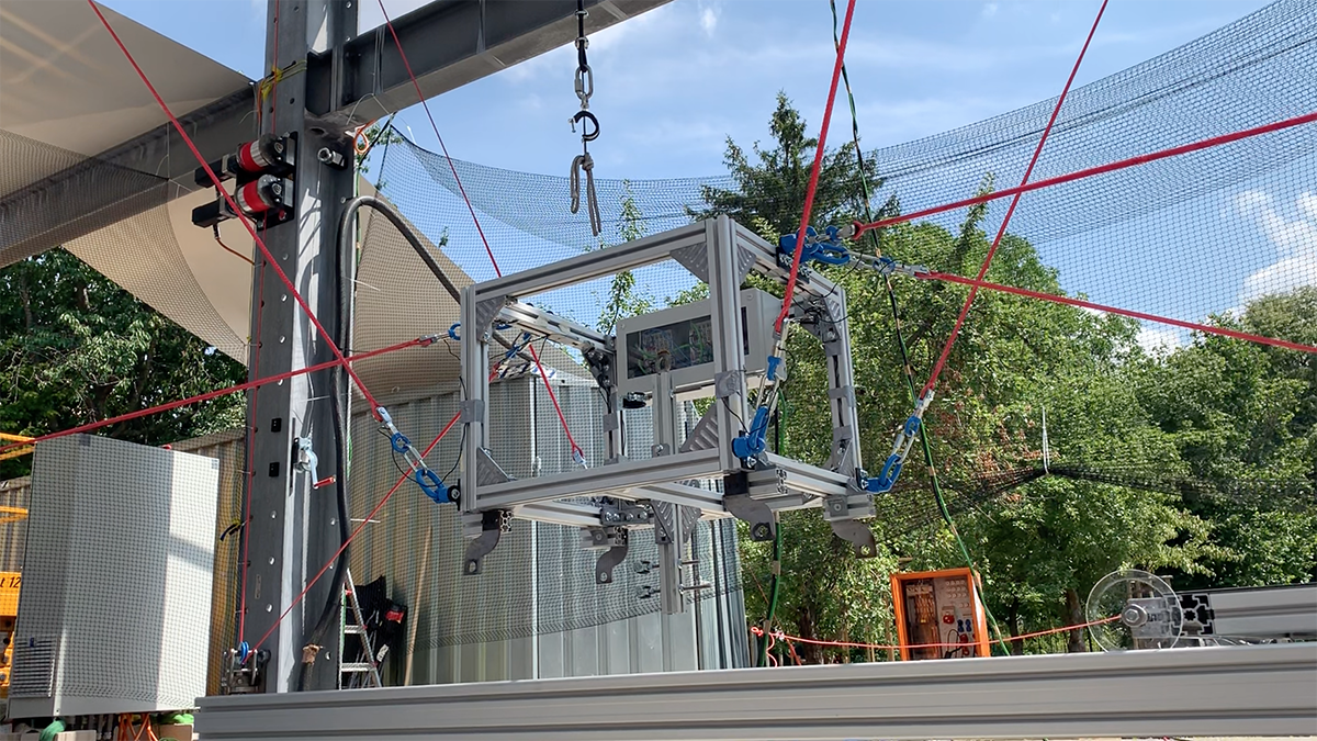 一个金属盒子形状的机器人被连接到各种红色电缆上，悬挂在几英尺高的空中。