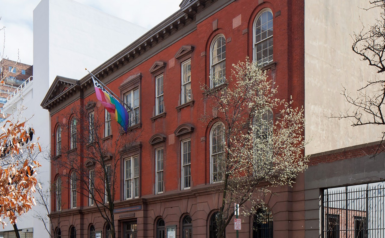 一块砖瓦房的一个透视照片在树排行的街道的。彩虹旗飞到建筑物的入口之上。