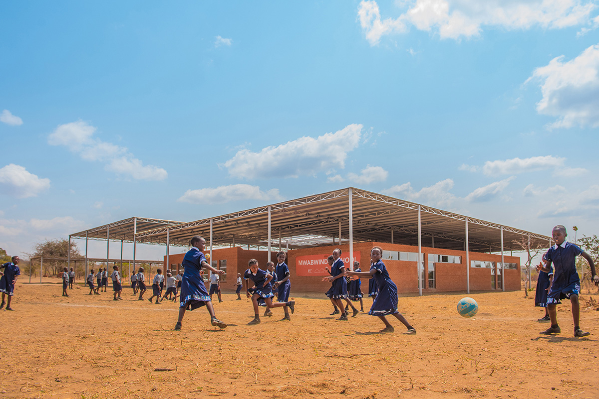 非洲孩子在砖砌学校门前踢足球，学校的牌子上写着Mwabwindo