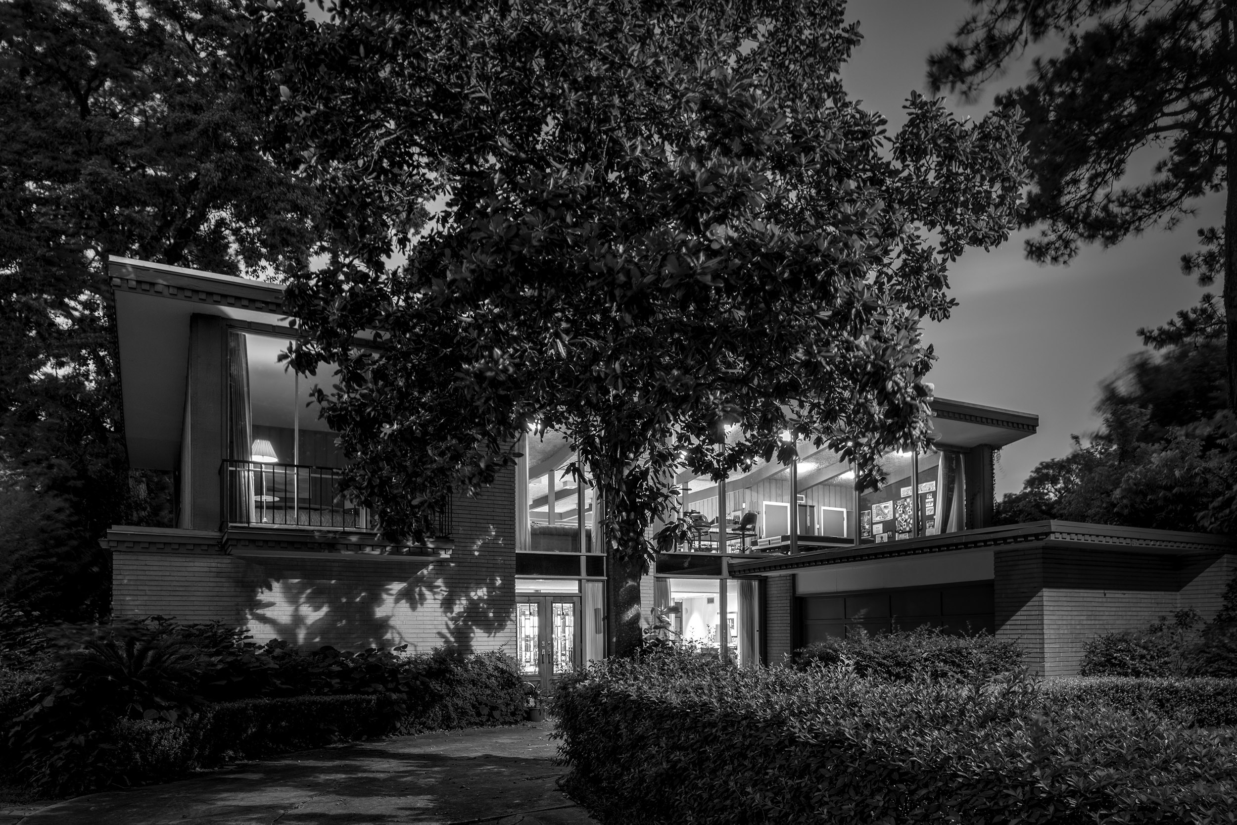 这张黑白照片描绘了休斯顿一座两层的世纪中期现代住宅，前景是一棵树