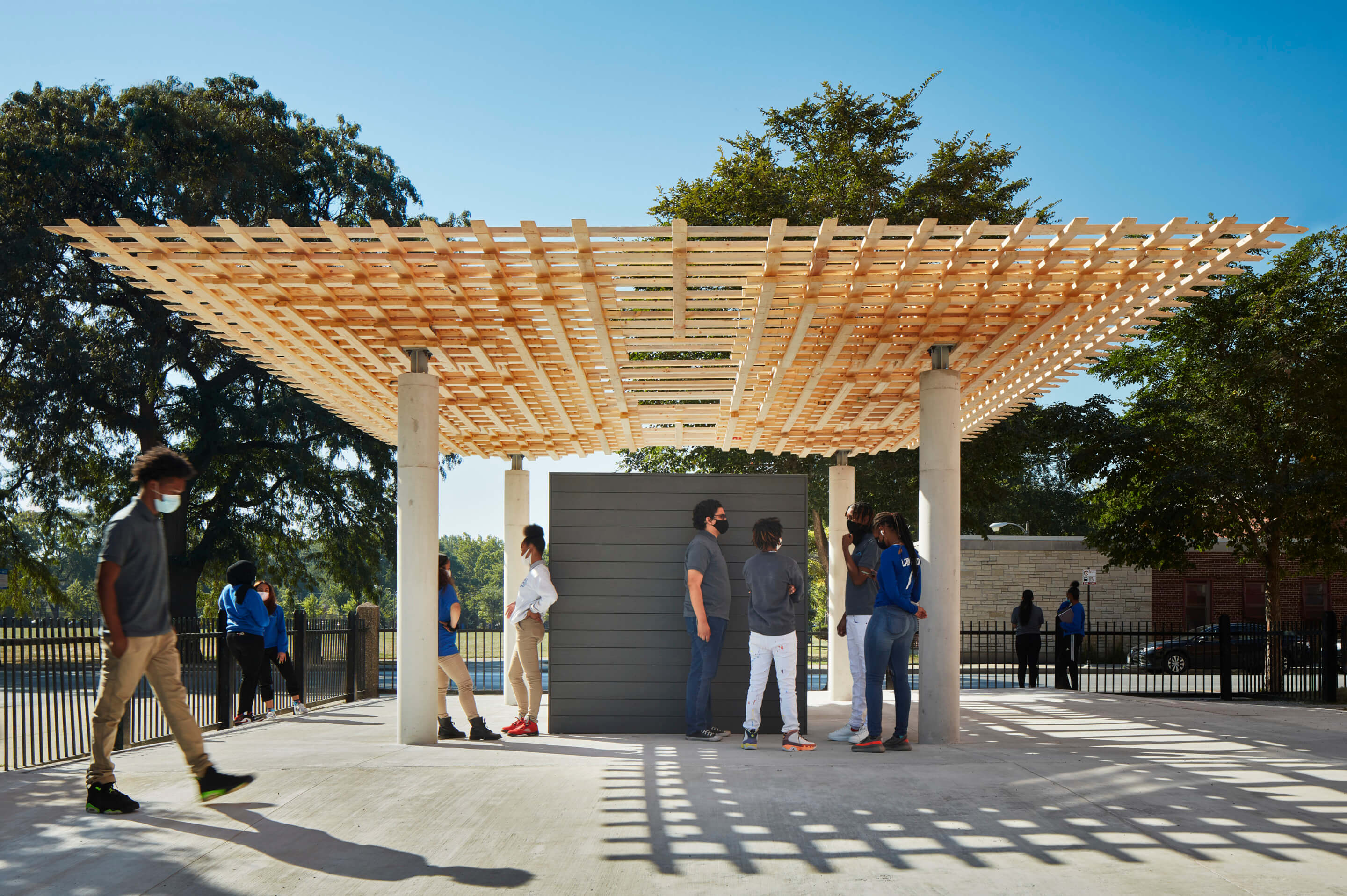 people gather beneath a sheltered outdoor pavilion with a timber roof structure