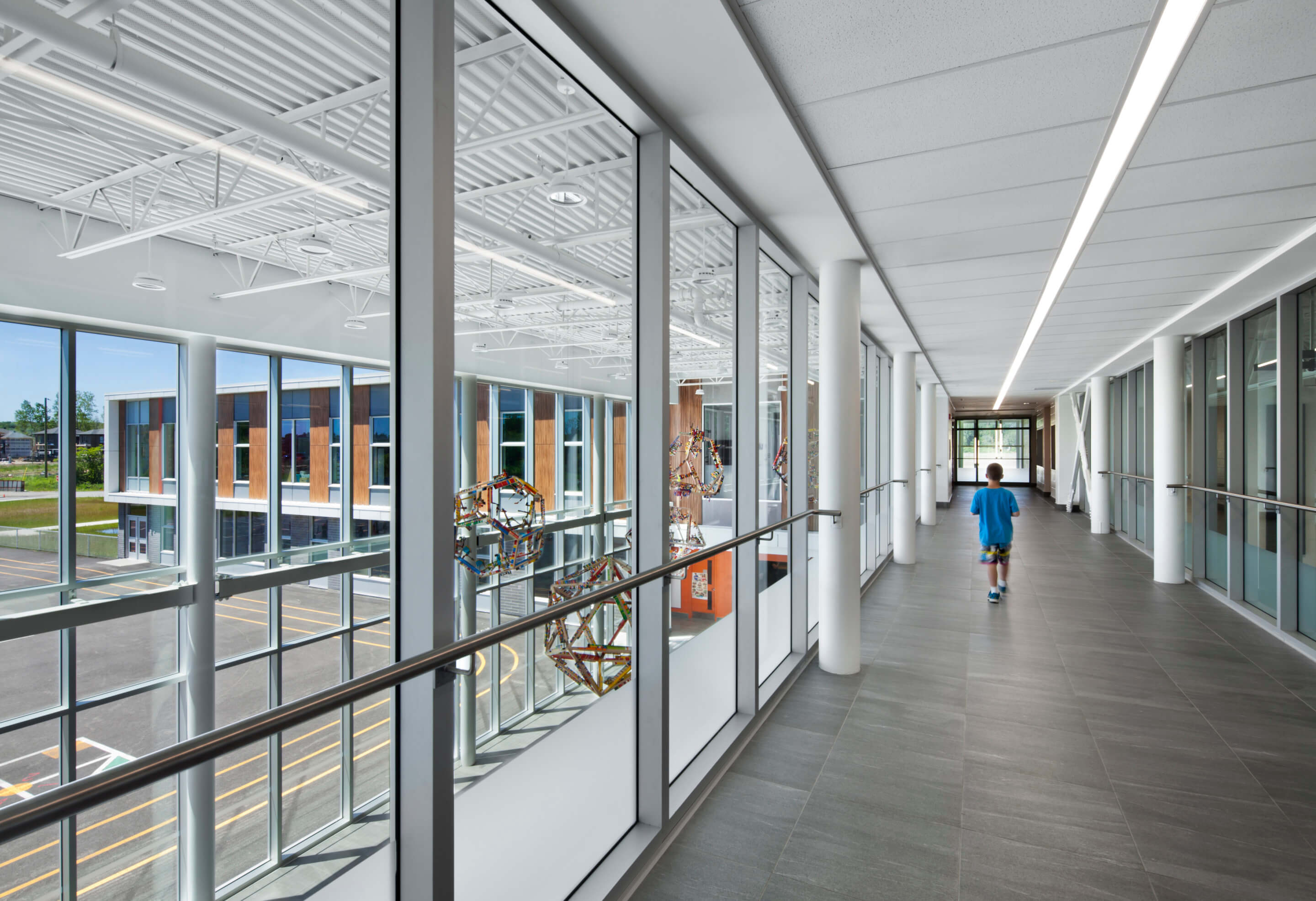 A long strip of light runs along the center of the ceiling of a glassed-in walkway that overlooks a two-story atrium